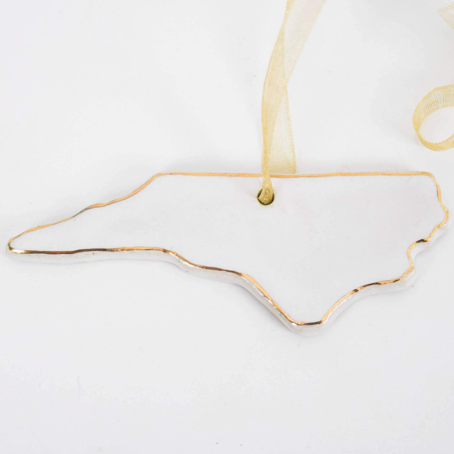 A hand-painted stoneware ornament in the shape of North Carolina with 22k gold edge detailing, hanging against a white background with a yellow ribbon.
