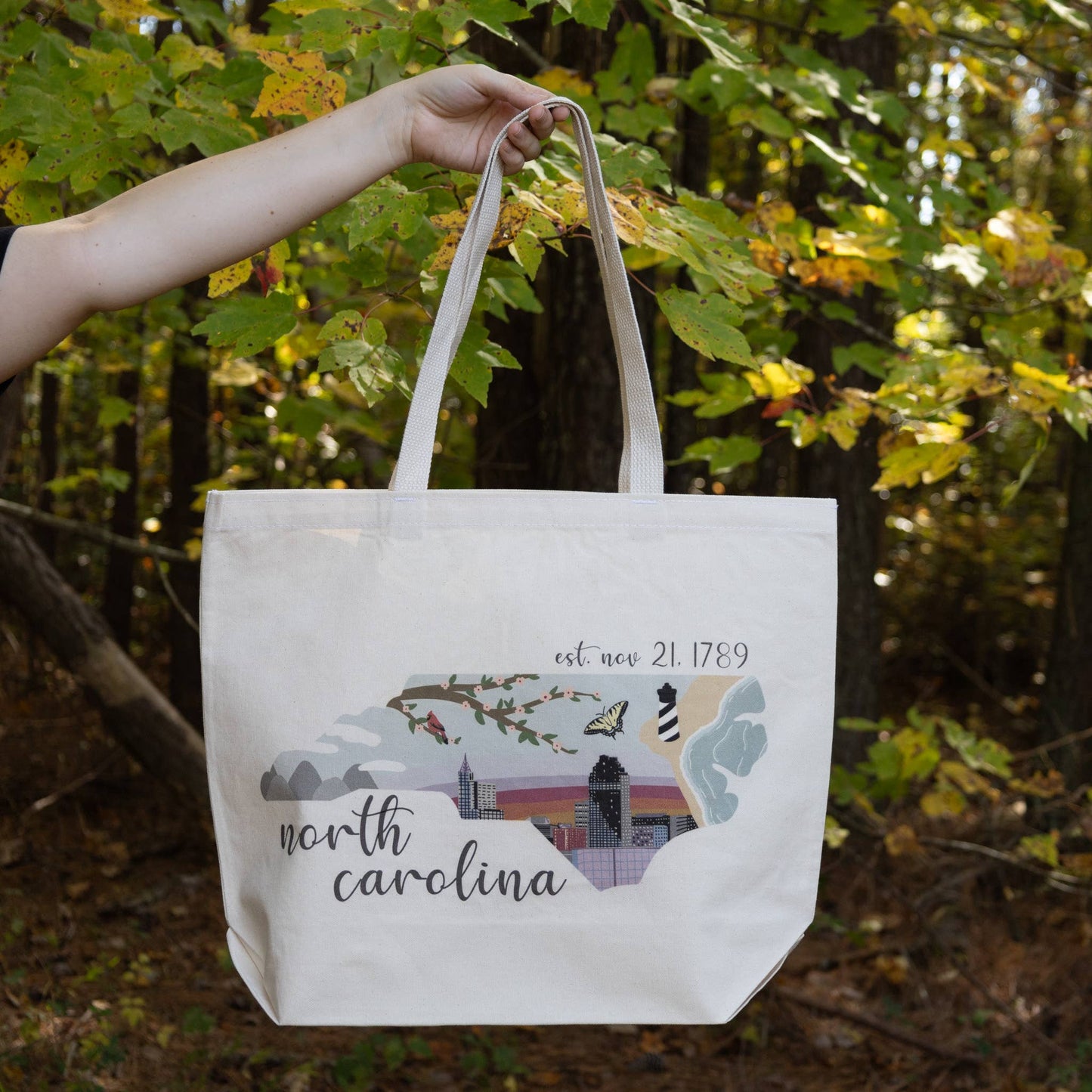 A beige jumbo tote bag with 'North Carolina' printed on the front, held in a hand with a background of trees.