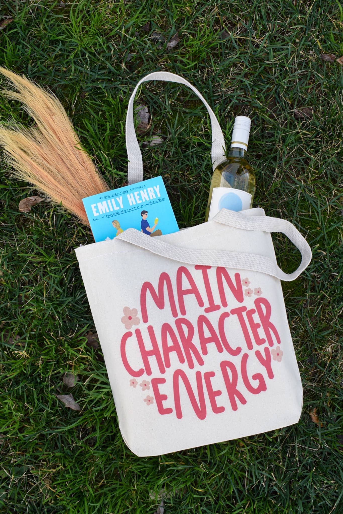 A pink cotton square tote bag with the text 'MAIN CHARACTER ENERGY' printed on the front, along with a book and a bottle of wine placed in it, set against a grassy background.