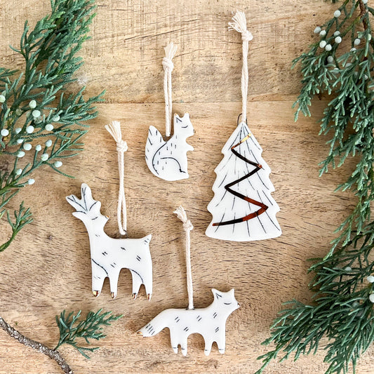 Set of five white mini ceramic woodland Christmas ornaments displayed on a wooden surface, including a tree, fox, deer, and squirrel shapes, each with simple black and gold line details and twine loops for hanging. Sprigs of greenery frame the scene.