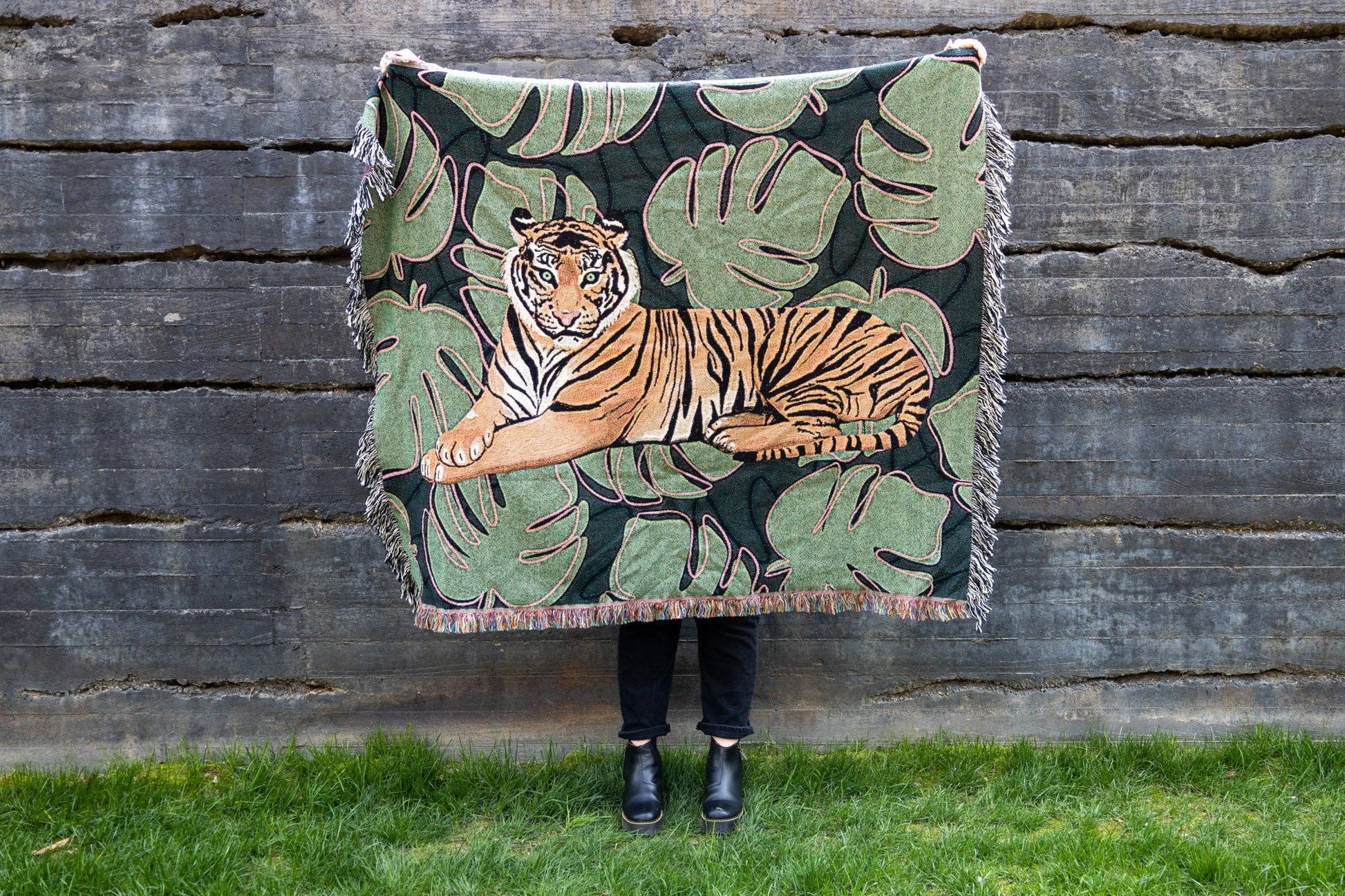 A green and brown Tiger Tapestry Throw Blanket with a central tiger design, surrounded by monstera leaves. A person's lower legs are visible from holding up the throw outside. the background is wooden.