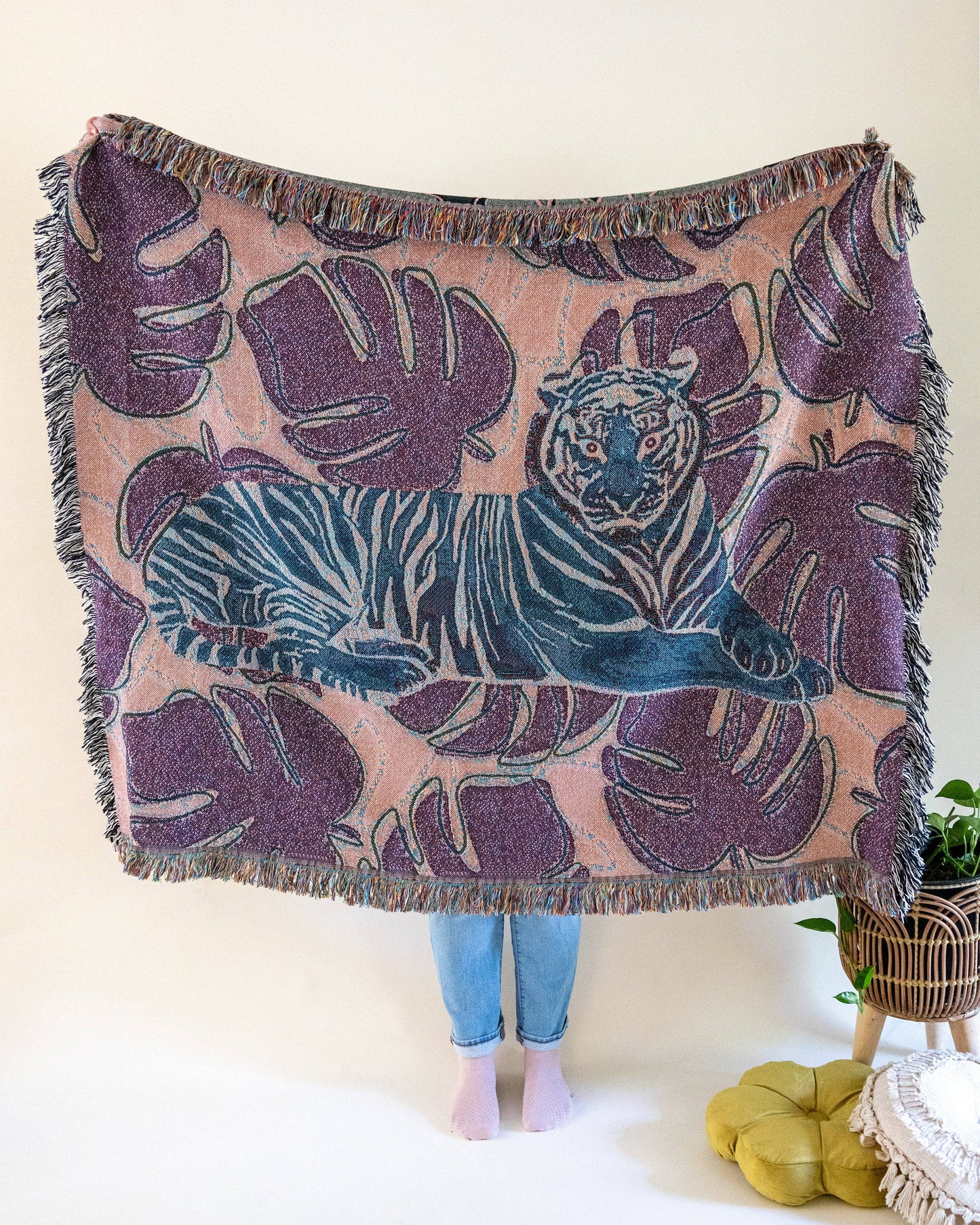 The reverse of the Tiger Tapestry Throw Blanket with a central tiger design, surrounded by monstera leaves.  The colors are salmon, blue, and purple. A person's lower legs are visible from holding up the throw.