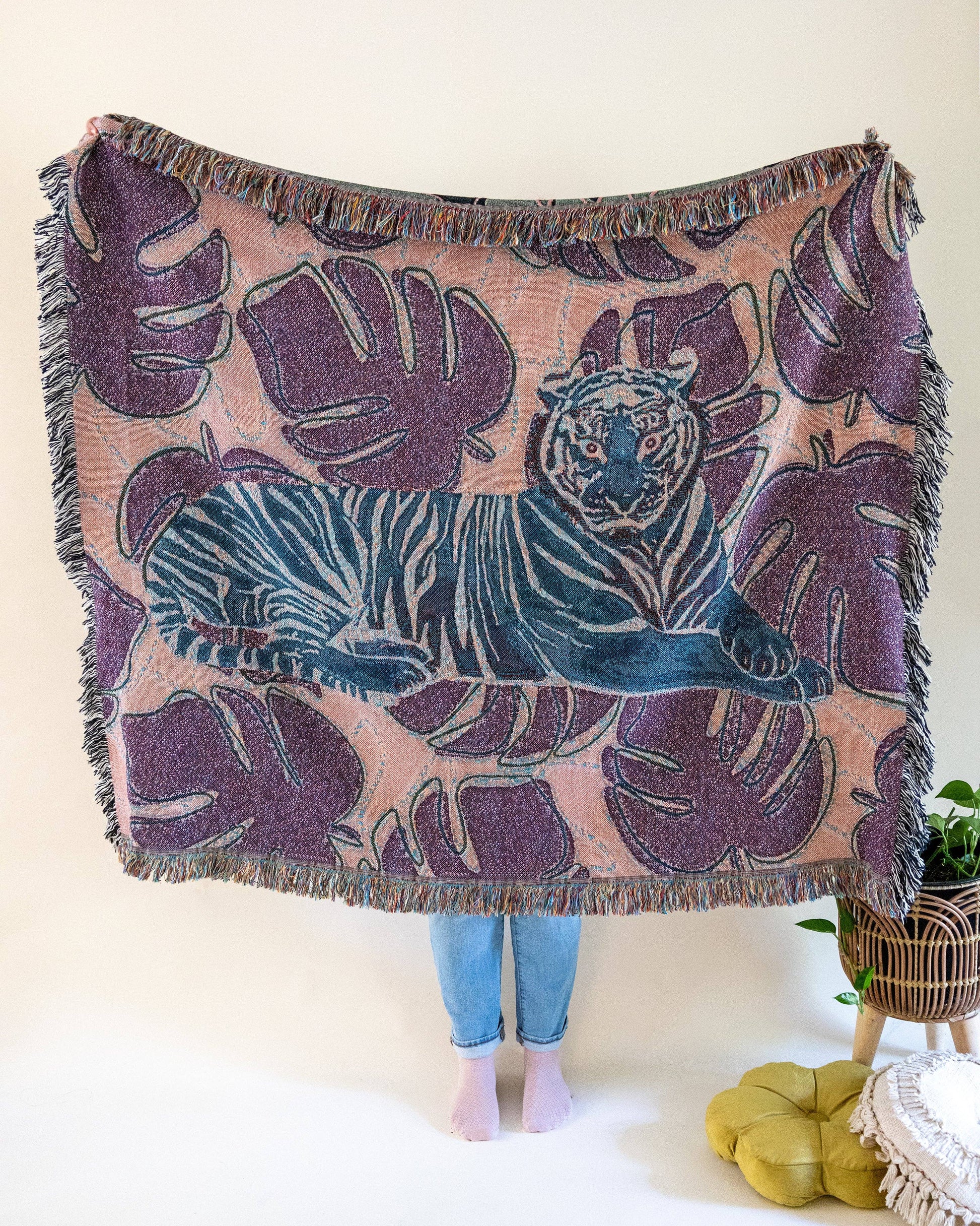 The reverse of the Tiger Tapestry Throw Blanket with a central tiger design, surrounded by monstera leaves.  The colors are salmon, blue, and purple. A person's lower legs are visible from holding up the throw.