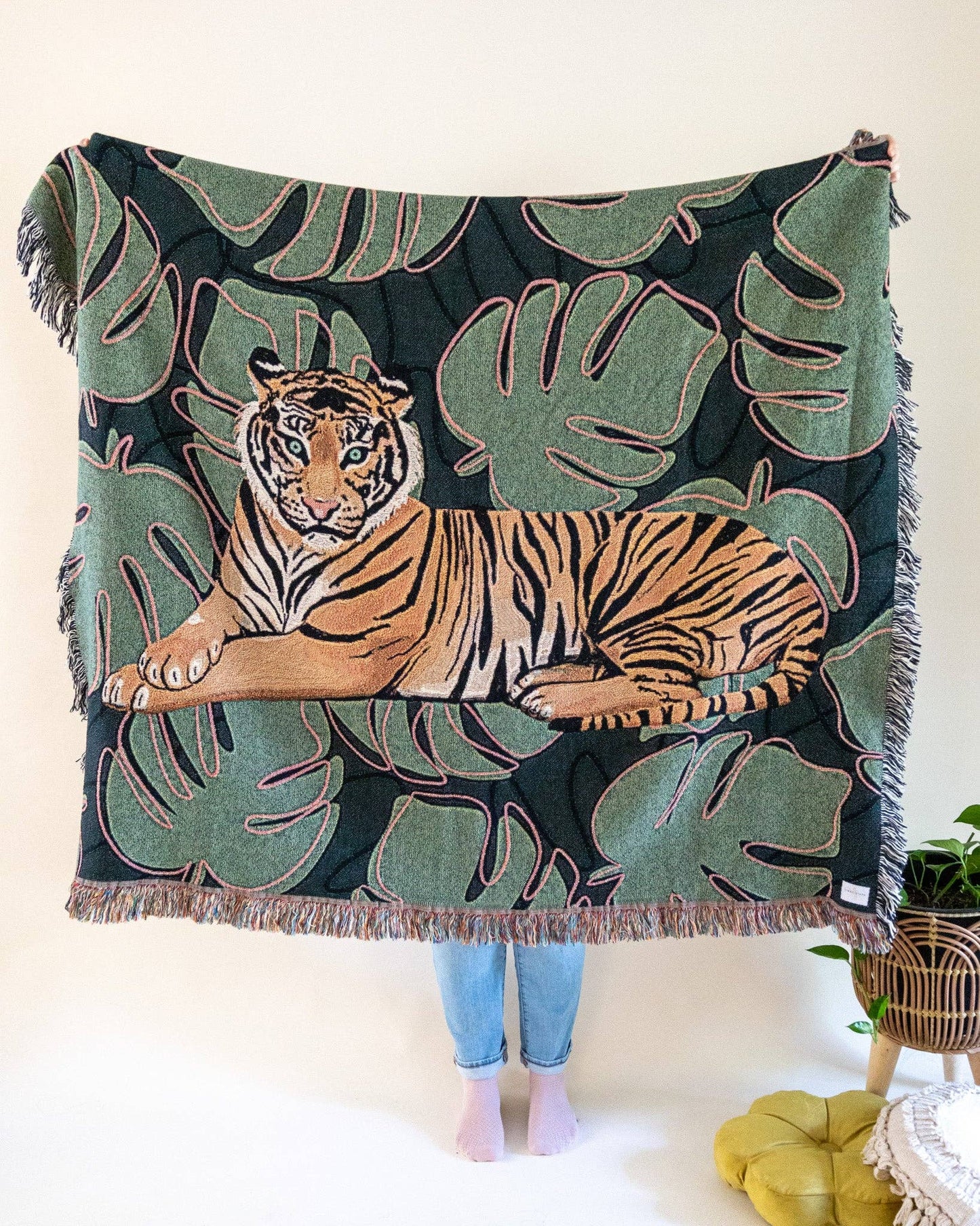 A green and brown Tiger Tapestry Throw Blanket with a central tiger design, surrounded by monstera leaves. A person's lower legs are visible from holding up the throw.