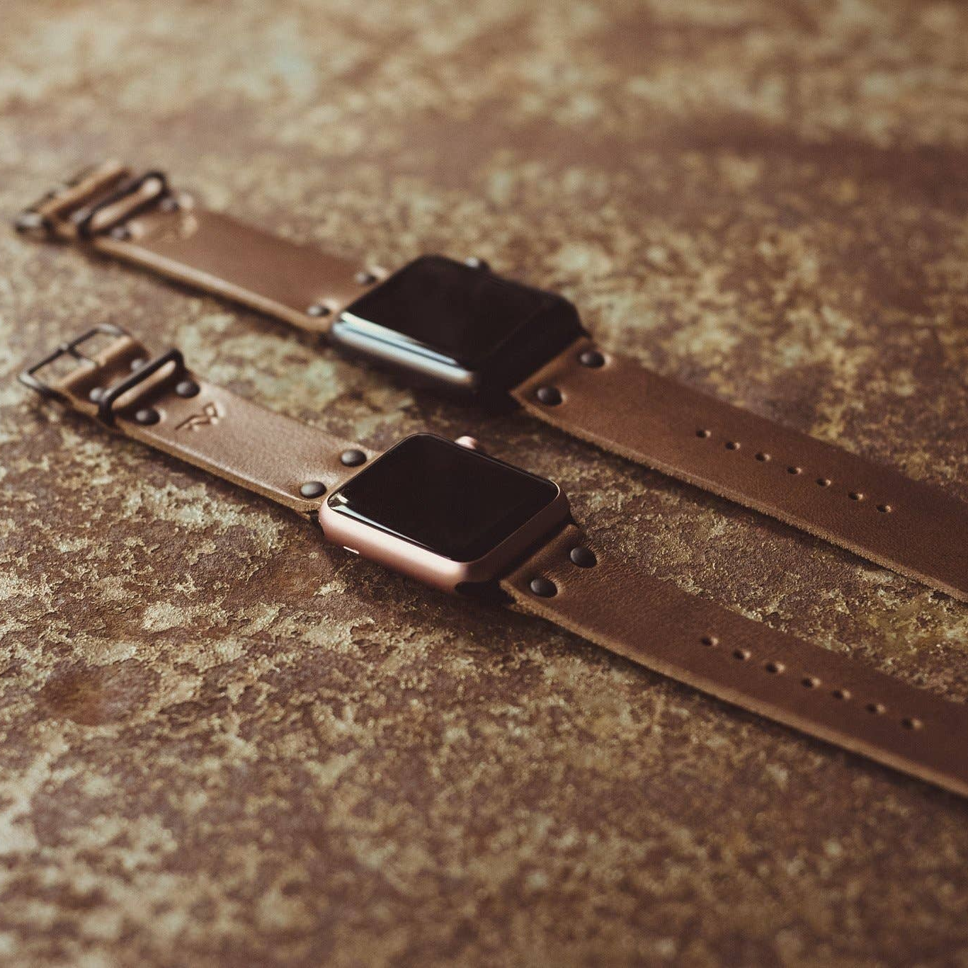 Two brown leather Apple Smart Watch bands, one laid out flat and the other on a curved surface, showcasing the watch band design and buckle.
