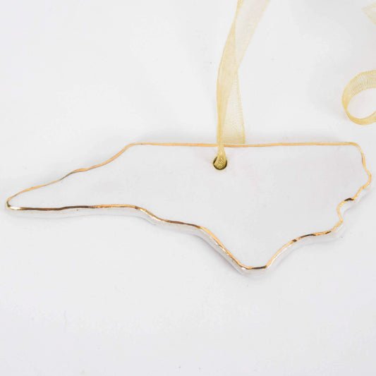 A hand-painted stoneware ornament in the shape of North Carolina with 22k gold edge detailing, hanging against a white background with a yellow ribbon.