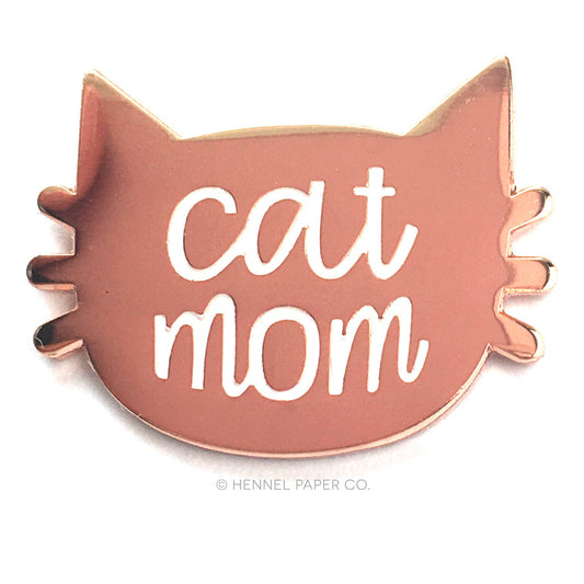 A rose gold metal lapel pin with the text 'CAT MOM' and a metal butterfly clasp, made by Hennel Paper Co.
